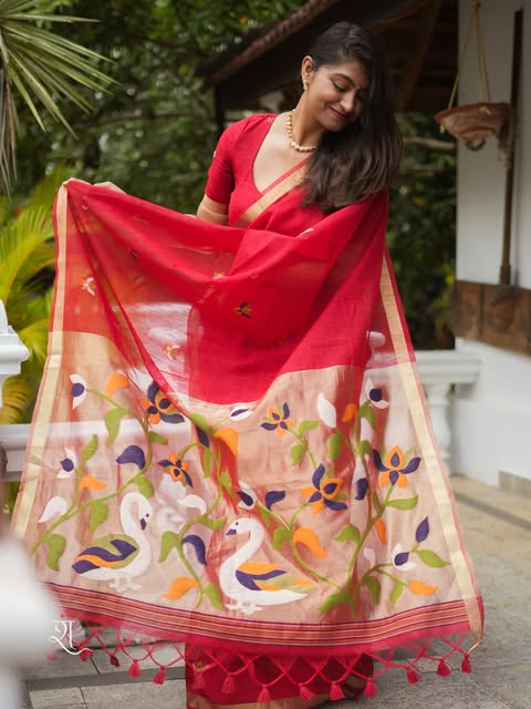 Hamsini by Ishmira, a red saree with an illustrated pallu, photographed in a leafy courtyard